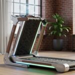 Sleek smart treadmill with wood-accented handrails and glowing LED strips set up in a modern loft with brick walls, tall windows, and potted plants.