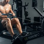 Muscular man using professional rowing machine in modern gym showing proper rowing technique with detailed performance display showing 2000m distance, 150 SPM stroke rate and workout timer