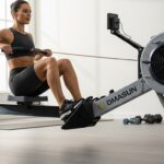 Athletic woman in black workout attire using a DMASUN rowing machine in a modern home gym setting, demonstrating proper rowing form with natural lighting from large windows and minimalist décor including dumbbells and plants